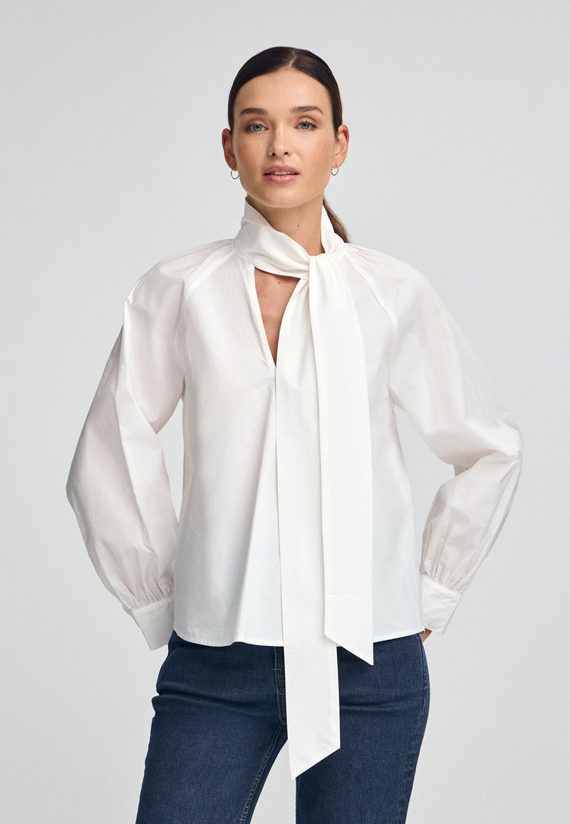 Blouse blanche en tissu lisse, avec un col à nouer, des épaules froncées et des manches longues avec des poignets élastiques. Portée avec un jean foncé.