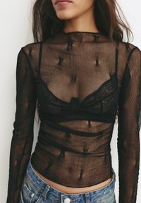 Sort gennemsigtig bluse med lange ærmer lavet af tekstureret stof, med høj hals og lagdelt design, båret over en sort blonde-bh.