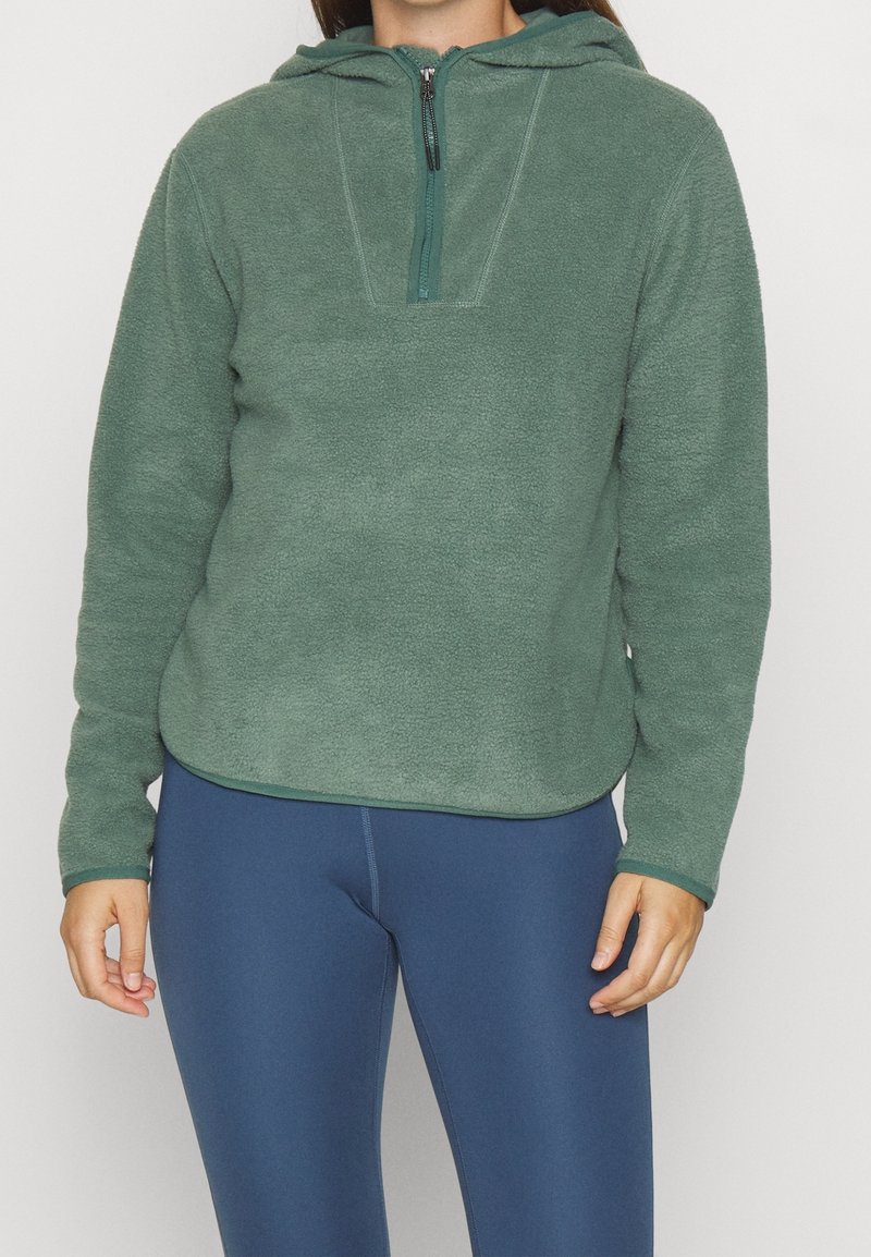 Felpa verde con zip a quarto davanti, cappuccio con cordino e una texture morbida. Abbinata a leggings blu navy.