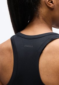 Κοντινή εικόνα του άνω μέρους της πλάτης ενός ατόμου που φοράει μαύρο αθλητικό τοπ τύπου racerback με τη λέξη «STRONGER» τυπωμένη με μικρά γράμματα.