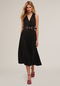 Schwarzes plissiertes Midikleid mit tiefem V-Ausschnitt und einem breiten schwarzen Gürtel mit goldenen Beschlägen. Kombiniert mit fuchsiafarbigen, hochhackigen Sandalen mit Riemchen.