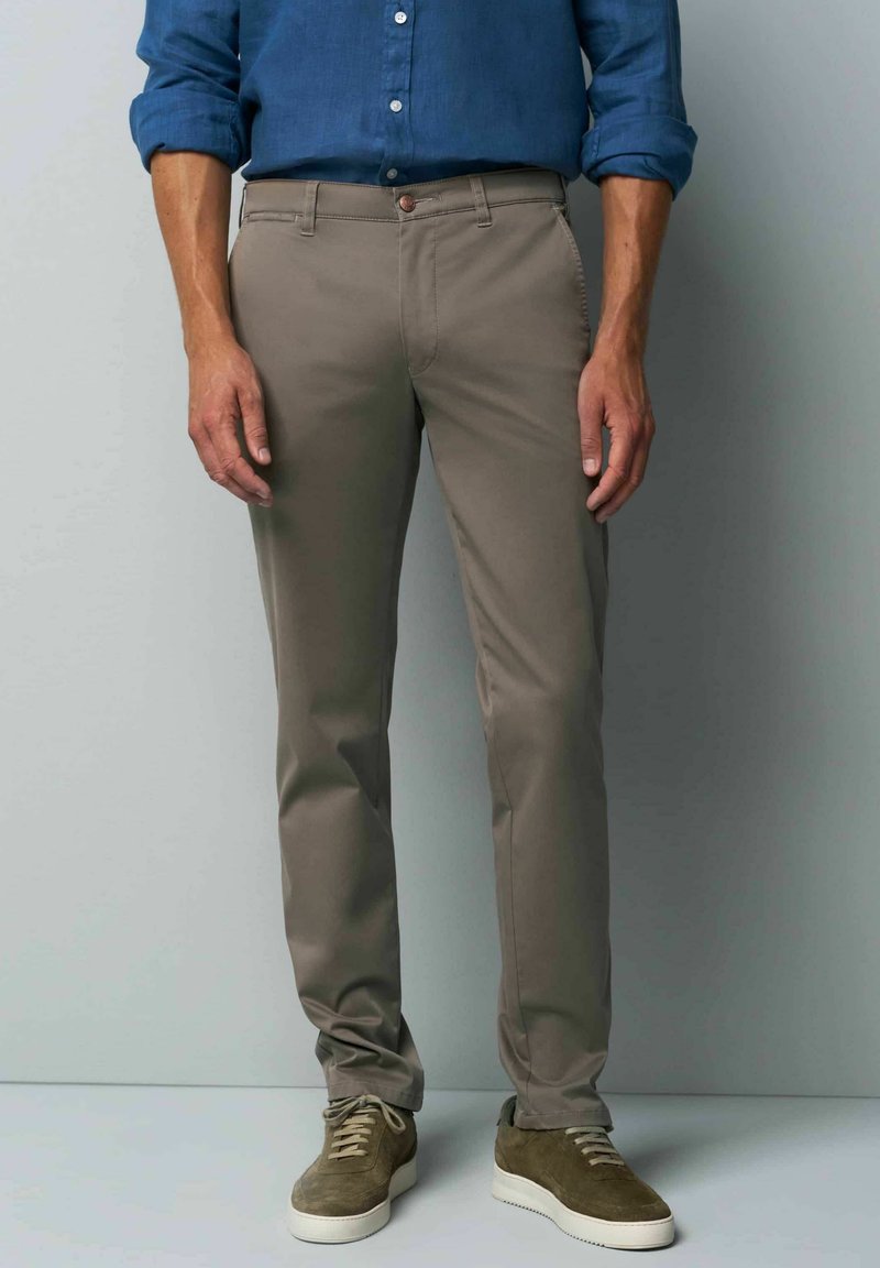 Beige chinos made of cotton blend, slim fit, straight legs, front pockets, and a small brown button detail at the waist. Paired with green sneakers.
