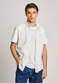 Camisa blanca de manga corta con rayas azul claro, llevada sobre una camiseta blanca. Combinada con jeans azules holgados. Diseño simple.