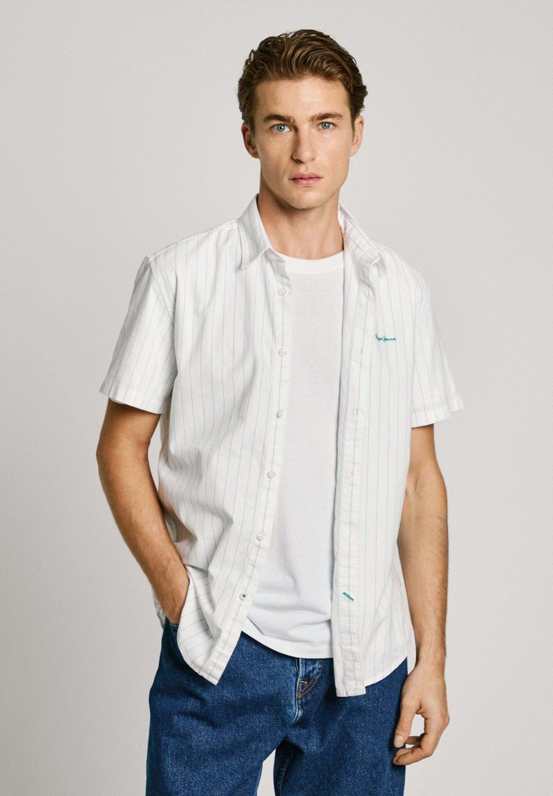 Camisa blanca de manga corta con rayas azul claro, llevada sobre una camiseta blanca. Combinada con jeans azules holgados. Diseño simple.