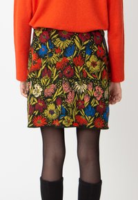 Bloemjesrok met felrode, blauwe en groene bloemen op een zwarte achtergrond. Draagt zwarte panty's en kniekousen.