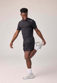 Maglietta sportiva nera con dettagli riflettenti, abbinata a pantaloni corti neri. Il modello esegue una posa di stretching, indossando calzini bianchi e sneakers grigie.