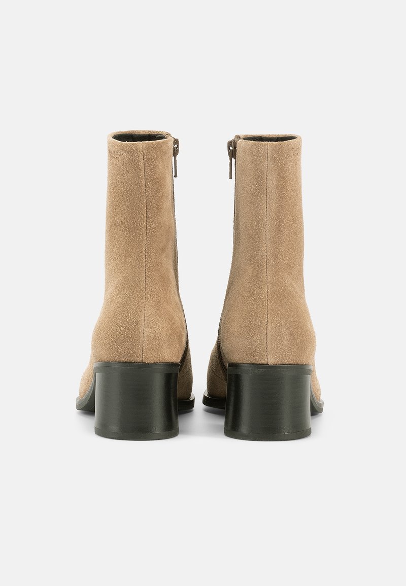 Beige ruskind ankelstøvler med et elegant design, der har en rundet tå og en sort blokhæl, samt en lynlås i siden til lukning.