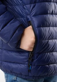 Giacca trapuntata blu navy con texture a trapuntatura, presenta una tasca laterale con zip e regolazioni con coulisse all'orlo per una vestibilità ottimale.