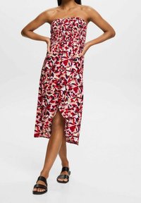 Blumenkleid ohne Träger mit gerafftem Oberteil, leuchtenden Rot- und Rosatönen, knielang, mit seitlichem Schlitz und leichtem Stoff. Schwarze Sandalen getragen.