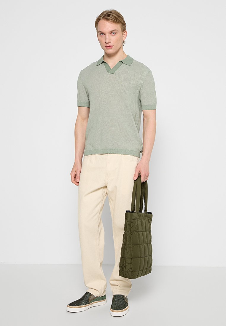 Hellgrünes Kurzarm-Polo mit gestrickter Textur, kombiniert mit hellbeigen Hosen und einer gesteppte olivgrünen Tragetasche. Sneaker mit grünen Netzakzenten.