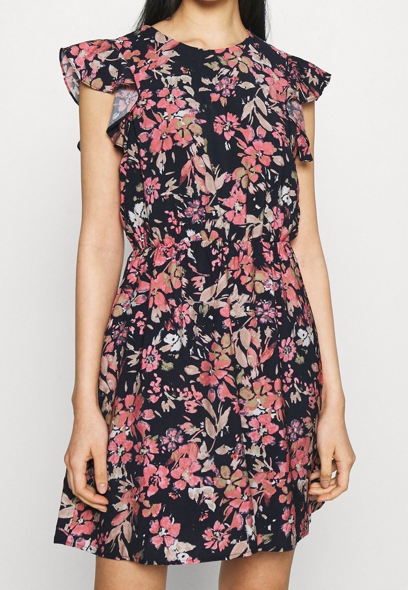 Femme portant une robe bleu marine sans manches avec des motifs floraux roses et beiges et des volants aux épaules, cintrée à la taille.