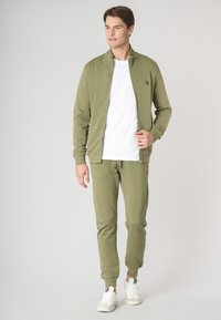 Tuta sportiva verde oliva composta da una giacca con zip e colletto alto e pantaloni jogger abbinati, entrambi realizzati in tessuto liscio, abbinati a sneakers bianche.