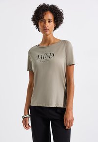 Oliivivärvi lühikeste varrukatega T-särk pehmest kangast, millel on must graafikaprindiga "MIND" tekst. Loodud vabaks istuvuseks, ümmargune kaelus.