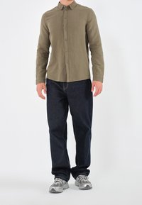 Camicia verde oliva a maniche lunghe, abbottonata, con colletto. Jeans blu scuro con cuciture visibili. Scarpe da ginnastica grigie con dettagli in rete.
