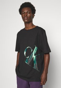 Black cotton t-shirt featuring a large, glossy green CK logo design on the front. Loose fit, crew neck with short sleeves.