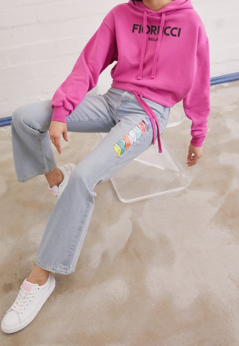 Sudadera con capucha rosa con logotipo negro, vaqueros acampanados azul claro con bordado de logotipo colorido, y zapatillas blancas con detalles en rosa.