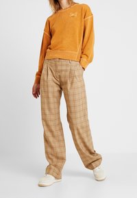 Orange sweatshirt med en texturerad yta och logotypmärke, kombinerad med beige rutiga byxor. Sneakers kompletterar outfiten med en avslappnad stil.