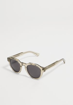 Lunettes de soleil rondes avec montures beiges transparentes et verres foncés posées sur une surface blanche.