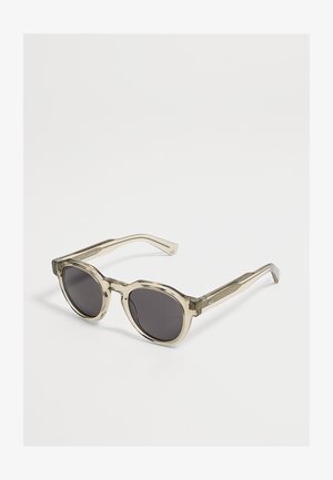 Lunettes de soleil rondes avec montures beiges transparentes et verres foncés posées sur une surface blanche.