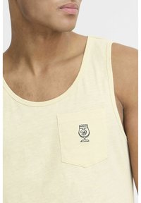 Heled kollane tank top, mis on valmistatud pehmest kangast, sisaldab rinnakotti, millel on tikitud klaasi disain. Lihtne, mugav lõige.