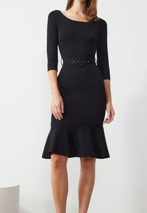 Vrouw gekleed in een nauwsluitende zwarte jurk met driekwartmouwen, een vierkante halslijn, een tailleriem en een uitlopende zoom boven de knieën.