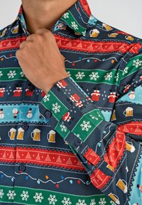 Man in een kerstthema knoopshirt met patronen van kerstmannetjes in schoorstenen, sneeuwvlokken, bierpullen, sneeuwmannen en kerstlichtjes.