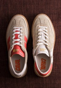 Paire de baskets en daim beige avec lacets blancs, accents rouges et marquage "CAVAL" sur les languettes et les semelles intérieures sur une surface texturée sombre.