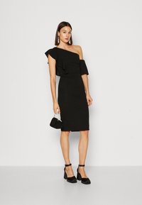 Rochie neagră cu o singură umăr, cu detalii cu volane, siluetă ajustată, până la genunchi, purtată împreună cu sandale cu toc negru și curea la gleznă și o geantă mică neagră.