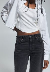 Weißes, gekürztes Shirt über einem weißen Tanktop, kombiniert mit schwarzen High-Waist-Jeans, die kupferfarbene Knopfdetails und kontrastierende Nähte aufweisen.