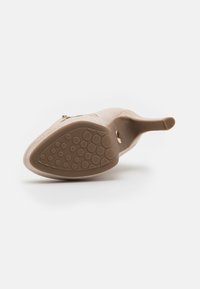 Chaussure à talon haut beige posée sur le côté, montrant une semelle texturée avec des motifs de grip ronds et un design à bout pointu.