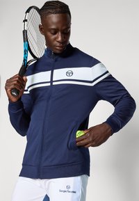 Marineblauw zip-up trainingsjack met witte streepaccenten. Heeft een logo op de borst en zijzakken, met een tennisbal zichtbaar in één zak.
