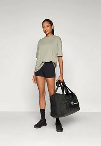 Light olive oversized t-shirt, black shorts, and black sneakers. Black duffel bag with white "Champion" logo. Smooth fabric and relaxed fit.