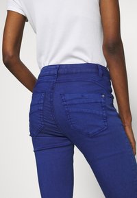 Höga blå jeans med en slät yta, som har bakfickor och en åtsittande form. Tyget har en lätt lyster.