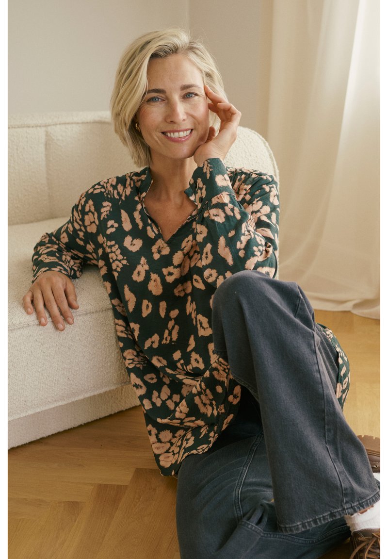 Femme souriante aux cheveux blonds courts, vêtue d'un chemisier à motifs et d'un jean foncé, assise sur un sol en bois, appuyée contre un canapé crème dans une pièce lumineuse.