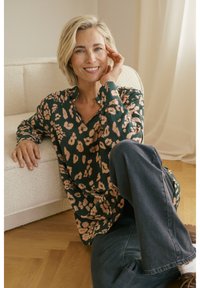 Femme souriante aux cheveux blonds courts, vêtue d'un chemisier à motifs et d'un jean foncé, assise sur un sol en bois, appuyée contre un canapé crème dans une pièce lumineuse.