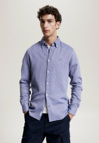 Camicia button-up a righe blu e bianche con colletto, maniche lunghe e un piccolo logo sul petto. Il tessuto ha un aspetto segnato.