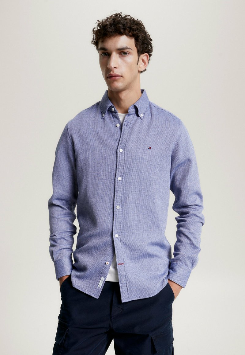 Camicia button-up a righe blu e bianche con colletto, maniche lunghe e un piccolo logo sul petto. Il tessuto ha un aspetto segnato.