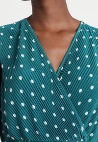 Teal wrap dress featuring a pleated texture with white polka dots. V-neck design with smooth, lightweight fabric and subtle sheen.