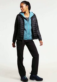 Svart pufferjakke over en turkise fleece, kombinert med svarte bukser og blå tursko. Har glidelås foran og elastiske mansjetter.