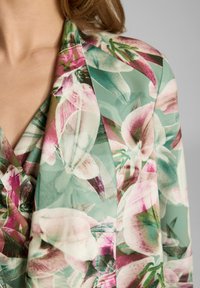 Blouse florale en tissu vert clair avec des motifs de fleurs roses et blanches, design à col en V et texture douce et transparente.