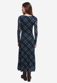 Nainen, jolla on yllä pitkähihaisen tummansinisen ruudullisen mekon, joka on kapea vyötäröltä. Hän seisoo selin, ja hänellä on jalassaan tummanruskeat keskipitkät saappaat.