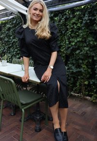 Rochie neagră cu mâneci bufante, decorată cu buline albe, cu o despicătură până la genunchi și material neted. Purtați-o cu pantofi loafer negri și bijuterii argintii.