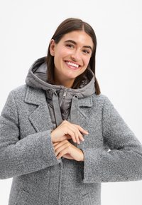 Cappotto grigio testurizzato con cappuccio imbottito grigio, dotato di chiusura con zip e cordini. Il cappotto presenta tasche laterali e una silhouette sagomata.