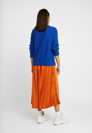 Kvinde stående med ryggen til iført en blå tekstureret sweater, orange midi-nederdel, hvide sokker og hvide chunky sneakers på en hvid baggrund.