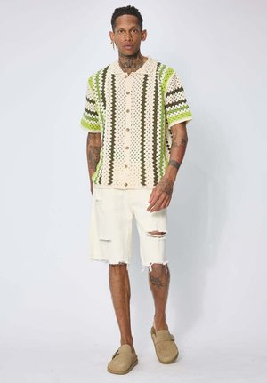 Homme tatoué portant une chemise courte à manches courtes en crochet rayée verte et crème, un short blanc déchiré et des chaussures beige à enfiler.