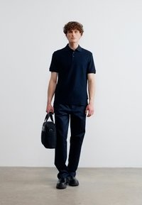 Νεαρός άνδρας που στέκεται σε λευκό τοίχο, φορώντας μαύρο πόλο, μαύρο παντελόνι, μαύρα παπούτσια, κρατώντας μαύρη τσάντα εργασίας στο δεξί χέρι.