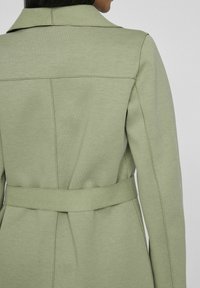Light green coat featuring a belted waist, structured collar, and smooth texture, with visible stitching and back seam details.