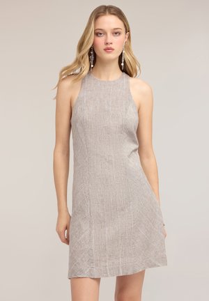 Mujer con el pelo largo y rubio que lleva un vestido sin mangas de color gris claro con textura y unos pendientes largos colgantes, de pie frente a un fondo liso.