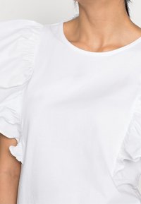 Top blanco de manga corta con mangas abullonadas y acampanadas, escote redondo y una textura de algodón suave. Sin patrones ni hardware visibles.