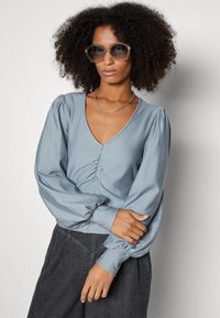 Blusa blu a maniche lunghe con scollatura a V e dettaglio arricciato sul davanti, realizzata in tessuto morbido; abbinata a pantaloni in denim scuro e occhiali da sole oversize.